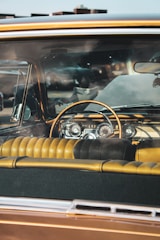 A vintage car interior viewed from the front windshield, featuring a classic dashboard with analog dials and an elegant steering wheel. The seats have a tufted leather design in shades of brown and yellow.