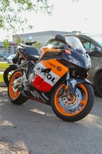 A sport motorcycle featuring a vibrant orange, black, and white color scheme with branding prominently displayed. It is parked on a street with grass and trees nearby, and a commercial building can be seen in the background.