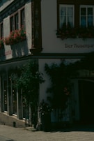 A charming, historic building with decorative flower boxes and lush greenery climbing the walls. The exterior features traditional architecture with arched windows and intricate woodwork. The sunlight casts shadows, creating a serene atmosphere.