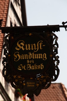 An ornate hanging sign with gold lettering in German, saying 'Kunsthandlung bei St Jakob'. The sign is framed with intricate ironwork and small decorative flowers. The background includes a blurred view of a building with traditional architecture, featuring a tiled roof and wooden elements.