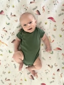 A baby wearing a green onesie lies on a white blanket decorated with illustrations of animals, mushrooms, butterflies, and leaves. The baby looks content, with a slight smile, and their arms and legs are slightly bent.