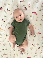 A baby wearing a green onesie lies on a white blanket decorated with illustrations of animals, mushrooms, butterflies, and leaves. The baby looks content, with a slight smile, and their arms and legs are slightly bent.