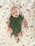A baby wearing a green onesie lies on a white blanket decorated with illustrations of animals, mushrooms, butterflies, and leaves. The baby looks content, with a slight smile, and their arms and legs are slightly bent.
