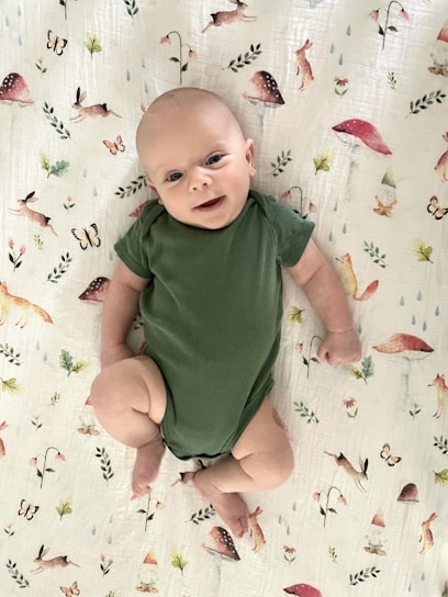 A baby wearing a green onesie lies on a white blanket decorated with illustrations of animals, mushrooms, butterflies, and leaves. The baby looks content, with a slight smile, and their arms and legs are slightly bent.