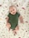 A baby wearing a green onesie lies on a white blanket decorated with illustrations of animals, mushrooms, butterflies, and leaves. The baby looks content, with a slight smile, and their arms and legs are slightly bent.