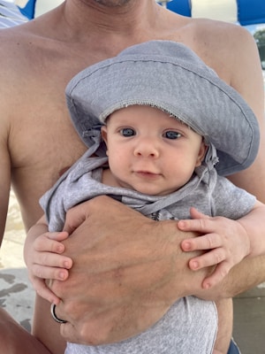 A baby wearing a gray sun hat, being held by an adult who is not wearing a shirt. The baby's hands are gently grasping the adult's hand. The background appears to be an outdoor setting with blue and white patterns, possibly an umbrella or tent.