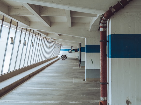 A parking garage with concrete pillars and a visible white car parked near the end. The garage features slanted glass windows on the left side and blue horizontal stripes painted on the walls. Red industrial pipes are visible attached to some pillars.