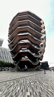 A modern architectural structure with multiple levels and walkways, featuring angular and geometric design elements. The building has a metallic and wooden exterior with large open spaces. It occupies a prominent position in a plaza surrounded by other contemporary buildings.