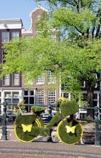 A bicycle creatively covered in green grass-like material is parked near a canal. Yellow butterflies adorn the wheels, and the bike is embellished with small flowers. Behind the bike, there are traditional European-style buildings and a large tree with vibrant green leaves.