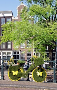 A bicycle creatively covered in green grass-like material is parked near a canal. Yellow butterflies adorn the wheels, and the bike is embellished with small flowers. Behind the bike, there are traditional European-style buildings and a large tree with vibrant green leaves.