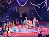 A vibrant circus performance featuring several elephants adorned with decorative blankets. Performers in colorful costumes, including dresses in blue, yellow, and red, are interacting with the elephants and each other. The scene is set on a circular stage with lights and ropes hanging from above, observed by an audience in the background.
