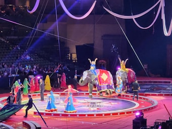 A vibrant circus performance featuring several elephants adorned with decorative blankets. Performers in colorful costumes, including dresses in blue, yellow, and red, are interacting with the elephants and each other. The scene is set on a circular stage with lights and ropes hanging from above, observed by an audience in the background.