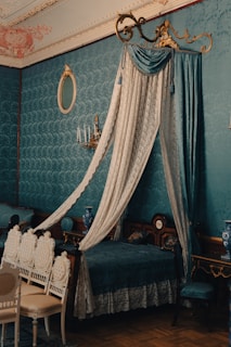 A richly decorated bedroom with vintage, upscale furnishings. A canopy bed draped in luxurious lace and velvet features prominently. Elegant chairs with intricate designs are arranged near the bed. The wall is adorned with ornate wallpaper, and a matching blue vase and a sconce with candles add to the room's classic ambiance. A gold-trimmed mirror finishes off the refined aesthetic.