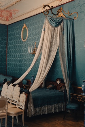 A richly decorated bedroom with vintage, upscale furnishings. A canopy bed draped in luxurious lace and velvet features prominently. Elegant chairs with intricate designs are arranged near the bed. The wall is adorned with ornate wallpaper, and a matching blue vase and a sconce with candles add to the room's classic ambiance. A gold-trimmed mirror finishes off the refined aesthetic.