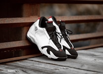 A pair of stylish black and white sneakers with red accents is placed on a wooden surface next to a rusty metal railing, creating a contrast between modern fashion and industrial elements.