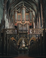 A grand pipe organ is situated at the focal point within a large cathedral interior, adorned with ornate architectural details. The organ is framed by pointed arches and intricate carvings, giving a sense of historical and artistic richness. The perspective is from the central aisle, flanked by wooden pews leading towards the altar.