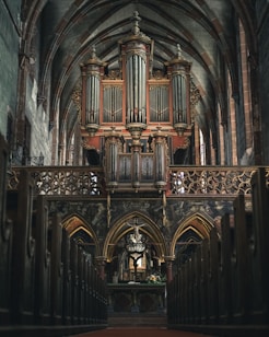 A grand pipe organ is situated at the focal point within a large cathedral interior, adorned with ornate architectural details. The organ is framed by pointed arches and intricate carvings, giving a sense of historical and artistic richness. The perspective is from the central aisle, flanked by wooden pews leading towards the altar.