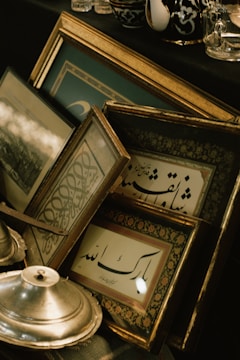 A collection of framed artworks with Arabic calligraphy leaning against each other. The frames have ornate golden details and are placed on a black surface along with a metallic dish and other decorative items. The setting suggests a vintage or classical theme with a focus on artistic expressions.