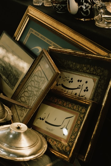 A collection of framed artworks with Arabic calligraphy leaning against each other. The frames have ornate golden details and are placed on a black surface along with a metallic dish and other decorative items. The setting suggests a vintage or classical theme with a focus on artistic expressions.