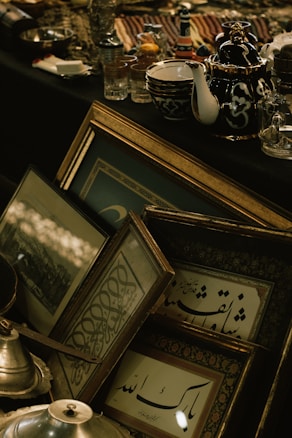 A collection of framed artworks and calligraphy pieces are arranged alongside a variety of ceramic and glass tableware. The frames are ornate, with golden and wooden finishes. A teapot and some bowls feature floral patterns, and there are numerous glass items like cups and jars scattered across a dark cloth surface.