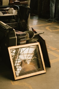 A collection of items is arranged on the floor including a vintage picture frame with a reflective surface, showing an abstract reflection of the surrounding area. Nearby are various objects like records, CDs, a pan, and other miscellaneous items. The scene is set at what appears to be a flea market or garage sale, with people standing in the background.