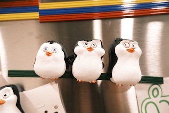 Three cartoon-style penguin magnets with big eyes and cheerful expressions are attached to a metallic surface. The background has colorful horizontal stripes, adding vibrancy to the scene.