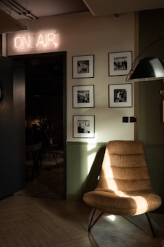 A cozy interior setting is depicted, featuring a modern leather chair positioned near a wall adorned with framed black-and-white photographs. Above an open doorway, a neon 'ON AIR' sign illuminates the space with a soft glow. The room is dimly lit, creating an intimate atmosphere, with light casting shadows on the chair.