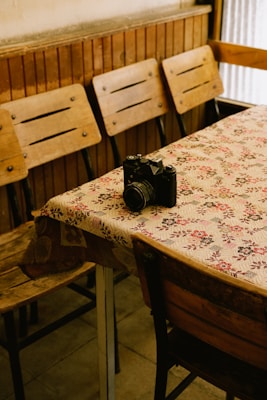 A vintage camera rests on a table covered with a floral-patterned cloth. The setting includes several wooden chairs with a rustic appearance. The room has a warm, nostalgic feel, emphasized by the wooden paneling and muted lighting.