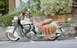 A vintage-style motorcycle with intricate leather saddlebags and chrome details is parked against a stone wall and green shrubbery. The bike has a mint green and cream color scheme with brown leather accents.