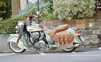 A vintage-style motorcycle with intricate leather saddlebags and chrome details is parked against a stone wall and green shrubbery. The bike has a mint green and cream color scheme with brown leather accents.