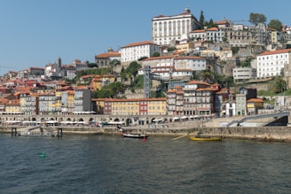 A picturesque coastal cityscape with colorful multi-story buildings stacked on a hillside, overlooking a river with boats. The architecture displays traditional styles with terracotta rooftops. The promenade along the river is bustling with outdoor cafes and people.
