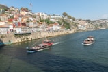 A scenic riverside view with two boats cruising along the water. The backdrop features colorful, historic buildings climbing up a hillside, with a mix of residential and commercial architecture. A bridge extends over the river in the background, and a crane is visible on the hill. The sunlit scene captures a blend of urban and natural beauty.