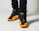 A close-up view of a person wearing black and yellow athletic sneakers. The shoes feature a distinctive brand logo on the tongue and a perforated design. The individual is wearing black pants, and the focus is primarily on the shoes.