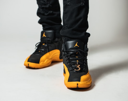 A close-up view of a person wearing black and yellow athletic sneakers. The shoes feature a distinctive brand logo on the tongue and a perforated design. The individual is wearing black pants, and the focus is primarily on the shoes.