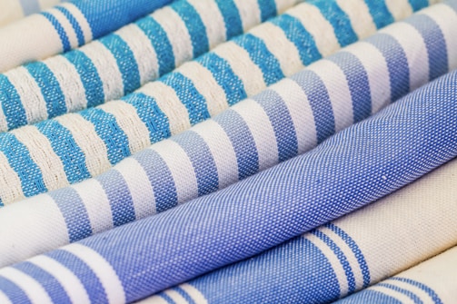 An array of rolled textiles with varying patterns, primarily featuring stripes in shades of blue and white. The fabric textures are visibly detailed, showcasing different weaving styles.