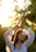 A woman in a white, flowy blouse is standing outdoors during sunset. She appears relaxed with her arms raised above her head. The sunlight creates a warm, golden glow around her, and the background is softly blurred with natural greenery.