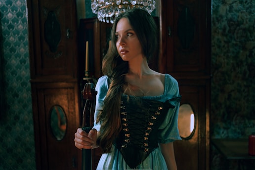 A woman wearing a historical-style dress with a corset stands indoors, holding an object that resembles a violin. The room appears to be dimly lit, with ornate wooden furniture and a chandelier overhead, emitting a warm, soft light. The setting suggests a vintage or historical theme.