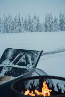 A snow-covered landscape with tall evergreen trees in the background. In the foreground, a snow shovel and a fire pit with flames provide a contrast to the wintry setting.