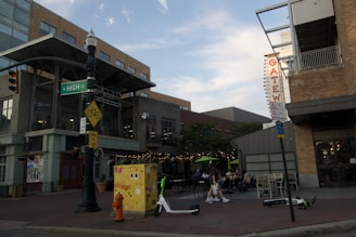 An urban street scene featuring a building with a modern design and a large 'Gateway' sign. Pedestrians are seen walking near outdoor seating areas with string lights and green umbrellas. A few electric scooters are scattered on the sidewalk. A street sign for 'N High St' is visible along with a bicycle sharing warning sign and a colorful utility box. The scene is set during daylight with a partly cloudy sky.