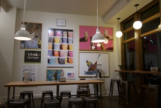 A cozy café interior featuring modern industrial design with metal stools, a wooden table, and pendant lights. The walls are adorned with colorful art and framed images, including ice cream cones and various food items. A prominent piece displays stacked ice cream pints, adding a quirky charm. The overall setting is illuminated warmly, creating a relaxed ambiance.