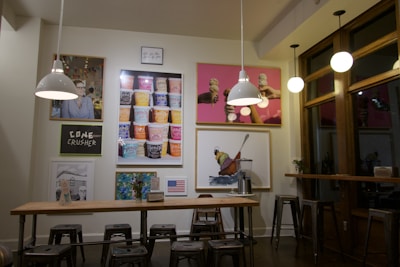 A cozy café interior featuring modern industrial design with metal stools, a wooden table, and pendant lights. The walls are adorned with colorful art and framed images, including ice cream cones and various food items. A prominent piece displays stacked ice cream pints, adding a quirky charm. The overall setting is illuminated warmly, creating a relaxed ambiance.