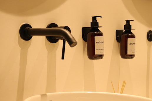 A modern bathroom setup featuring two wall-mounted soap dispensers and a sleek, black faucet above a white sink. The dispensers are labeled 'volumen' and set against a cream-colored wall. Below the dispensers, a set of wooden sticks is partially visible, suggesting a minimalist style.