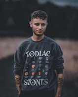 A person stands outdoors wearing a dark sweatshirt featuring various colorful gemstone illustrations labeled with zodiac names. The background is blurred, highlighting the figure. The person's arms are visible with tattoos, and they have curly hair.