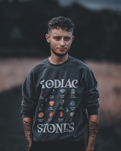 A person stands outdoors wearing a dark sweatshirt featuring various colorful gemstone illustrations labeled with zodiac names. The background is blurred, highlighting the figure. The person's arms are visible with tattoos, and they have curly hair.