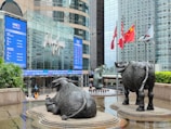 A modern urban setting featuring a large commercial building with reflective glass windows. Prominent in the foreground are two bronze bull sculptures, symbolizing economic strength and power. Surrounding the sculptures are several national flags on flagpoles, and lush green plants line the walkway. People are visible walking near the entrance of the building, evoking a sense of busy city life.