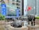 A modern urban setting featuring a large commercial building with reflective glass windows. Prominent in the foreground are two bronze bull sculptures, symbolizing economic strength and power. Surrounding the sculptures are several national flags on flagpoles, and lush green plants line the walkway. People are visible walking near the entrance of the building, evoking a sense of busy city life.