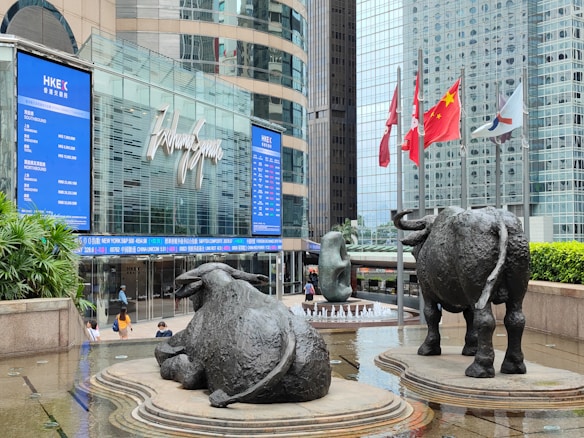 A modern urban setting featuring a large commercial building with reflective glass windows. Prominent in the foreground are two bronze bull sculptures, symbolizing economic strength and power. Surrounding the sculptures are several national flags on flagpoles, and lush green plants line the walkway. People are visible walking near the entrance of the building, evoking a sense of busy city life.