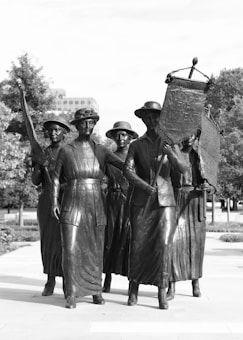 A group of statues depicting five women dressed in early 20th-century clothing. They are holding banners, one of which reads 'Votes for Women.' The scene is set outdoors, with trees and buildings in the background.