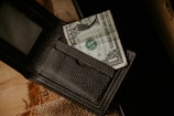 A textured black wallet partially open with a one-dollar bill protruding from one of the compartments. The wallet is placed on a surface that seems to be wooden and has a coarse woven mat beneath it.