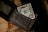 A textured black wallet partially open with a one-dollar bill protruding from one of the compartments. The wallet is placed on a surface that seems to be wooden and has a coarse woven mat beneath it.